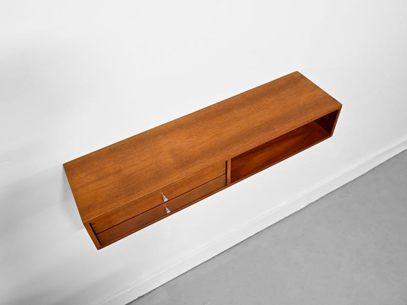 May include: A rectangular, wall-mounted wooden shelf with a warm brown finish. The shelf features two drawers with silver handles on the left and an open compartment on the right. The shelf is mounted on a white wall.