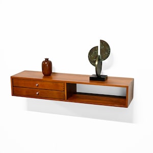 May include: A floating wooden shelf with two drawers and an open compartment. A small brown vase and a bronze sculpture with a black base are displayed on the shelf. The shelf is made of wood and is mounted on a white wall.
