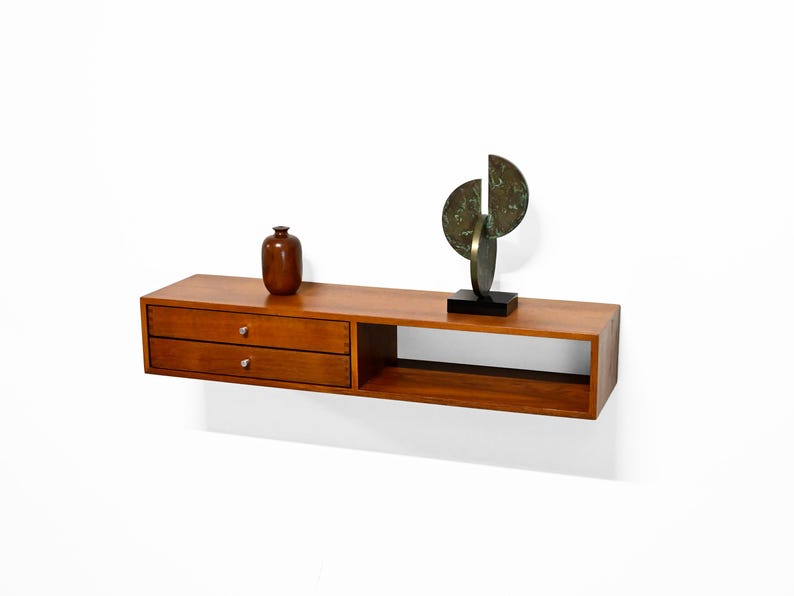 May include: A floating wooden shelf with two drawers and an open compartment. A small brown vase and a bronze sculpture with a black base are displayed on the shelf. The shelf is made of wood and is mounted on a white wall.