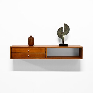 May include: A floating wooden shelf with two drawers and a lower open section. A brown vase and a bronze sculpture with a black base are displayed on the shelf. The shelf is mounted on a white wall.