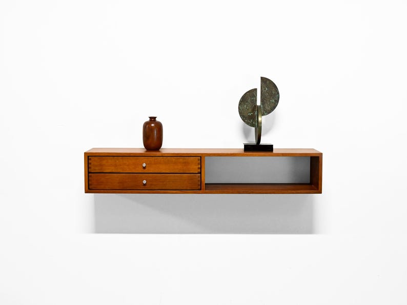 May include: A floating wooden shelf with two drawers and a lower open section. A brown vase and a bronze sculpture with a black base are displayed on the shelf. The shelf is mounted on a white wall.