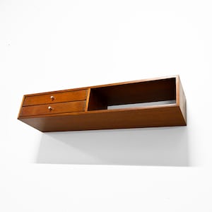 May include: A floating wooden shelf with two drawers and a cubby. The shelf is a warm brown color with gold-colored knobs on the drawers. The shelf is mounted on a white wall.