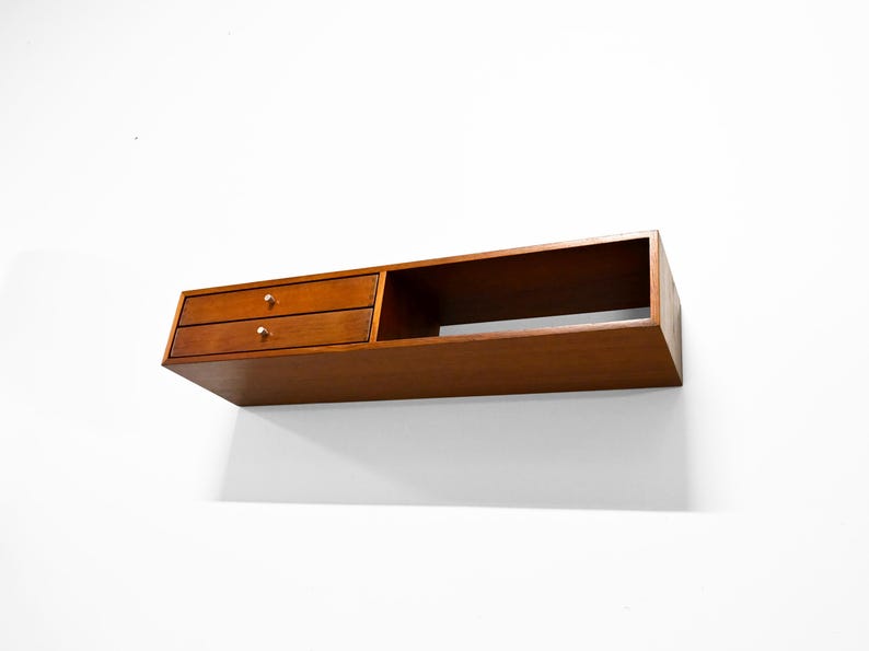 May include: A floating wooden shelf with two drawers and a cubby. The shelf is a warm brown color with gold-colored knobs on the drawers. The shelf is mounted on a white wall.