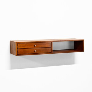May include: A wall-mounted wooden shelf with a warm brown finish. It features two drawers with silver knobs on the left and an open compartment on the right. The shelf is designed for storage and display.