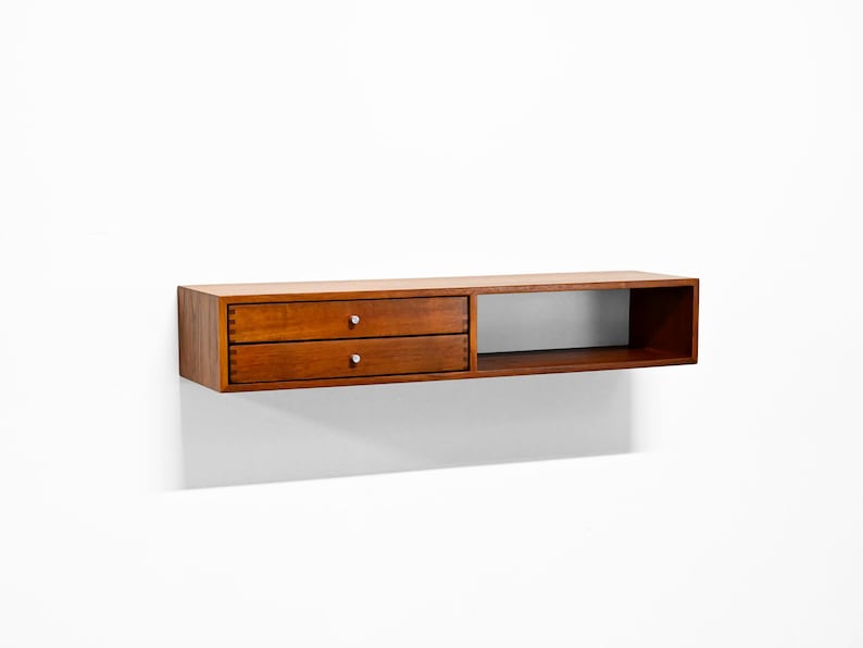 May include: A wall-mounted wooden shelf with a warm brown finish. It features two drawers with silver knobs on the left and an open compartment on the right. The shelf is designed for storage and display.