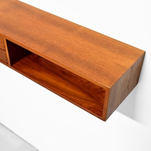 May include: A floating wooden shelf with a warm, reddish-brown finish. The shelf features two small drawers with silver handles on the left and an open compartment on the right. The shelf is mounted on a white wall.