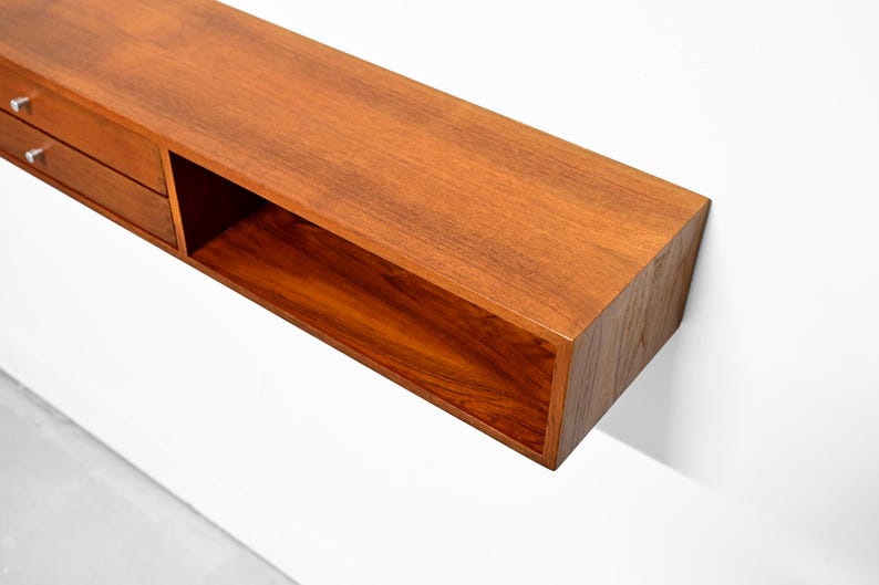 May include: A floating wooden shelf with a warm, reddish-brown finish. The shelf features two small drawers with silver handles on the left and an open compartment on the right. The shelf is mounted on a white wall.