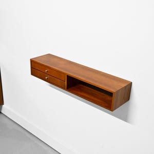 May include: A floating wooden shelf with two drawers and an open compartment. The shelf is a warm brown color and has silver drawer pulls. It is mounted on a white wall, showcasing a minimalist design.