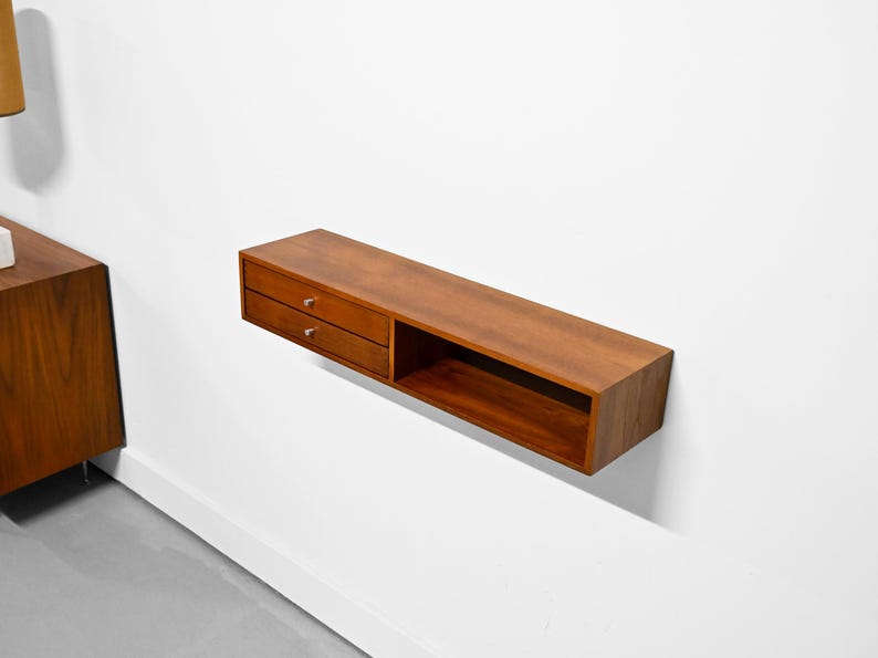 May include: A floating wooden shelf with two drawers and an open compartment. The shelf is a warm brown color and has silver drawer pulls. It is mounted on a white wall, showcasing a minimalist design.