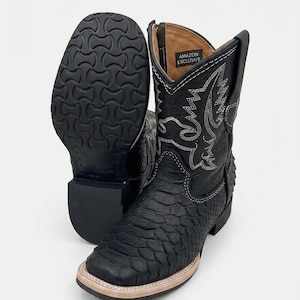 May include: Black cowboy boots with a textured, snake-like pattern. The boots have a square toe, decorative stitching, and a light-colored sole. The sole of one boot is visible, showing a patterned tread. The boots are labeled "AMAZON EXCLUSIVE".
