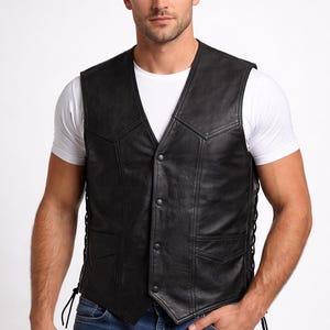 May include: Black leather vest with a V-neck, button closure, and side lacing. The vest has two front pockets and is worn over a white t-shirt and blue jeans. The vest is a classic biker style.