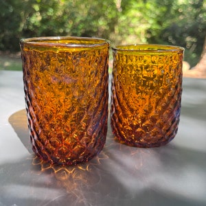 May include: Two amber glass tumblers with a textured, diamond-shaped pattern. The glasses are empty and sitting on a white surface.