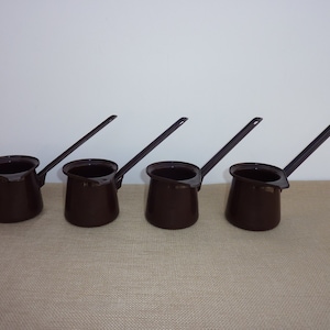 May include: Four brown metal coffee pots with long handles. The pots are lined up in a row on a brown surface.
