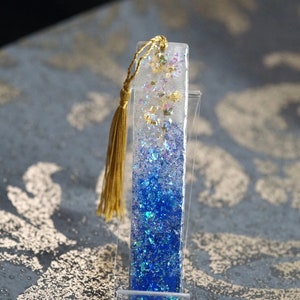 Könnte beinhalten: Rechteckiger Lesezeichen aus Harz mit blauem Glitzerverlauf, Goldblattakzenten und einer goldenen Quaste. Das Lesezeichen wird in einem transparenten Acrylständer präsentiert. Ein einzigartiges Accessoire für Bücherliebhaber.