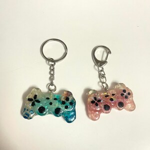 Może przedstawiać: Dwa breloczki do kluczy z miniaturowymi kontrolerami do gier. Jeden kontroler jest niebiesko-zielony z czarnymi akcentami, drugi r&oacute;żowy z czarnymi akcentami. Oba mają srebrne k&oacute;łka i łańcuszki.