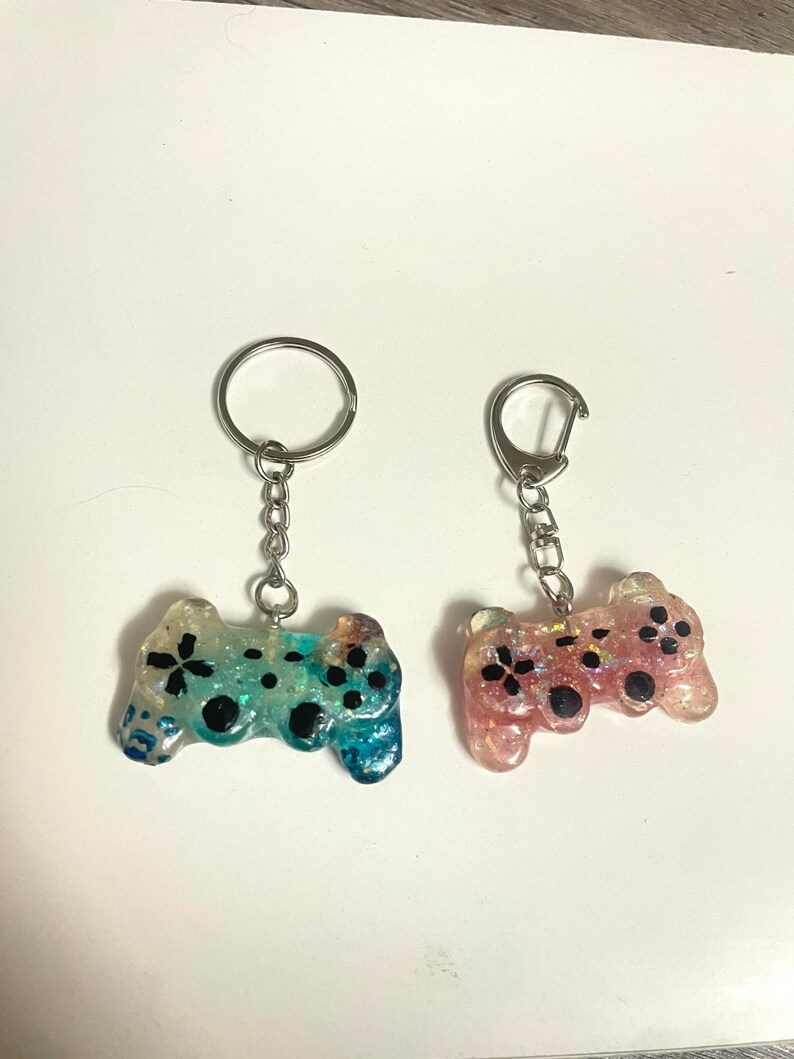 Może przedstawiać: Dwa breloczki do kluczy z miniaturowymi kontrolerami do gier. Jeden kontroler jest niebiesko-zielony z czarnymi akcentami, drugi r&oacute;żowy z czarnymi akcentami. Oba mają srebrne k&oacute;łka i łańcuszki.