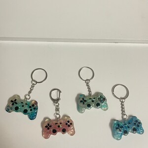 Może przedstawiać: Cztery breloczki do kluczy z żywicy w kształcie kontroler&oacute;w do gier wideo. Każdy breloczek ma srebrny pierścień i łańcuszek. Kontrolery są w odcieniach niebieskiego, r&oacute;żowego i zielonego, z czarnymi detalami guzik&oacute;w i brokatem.