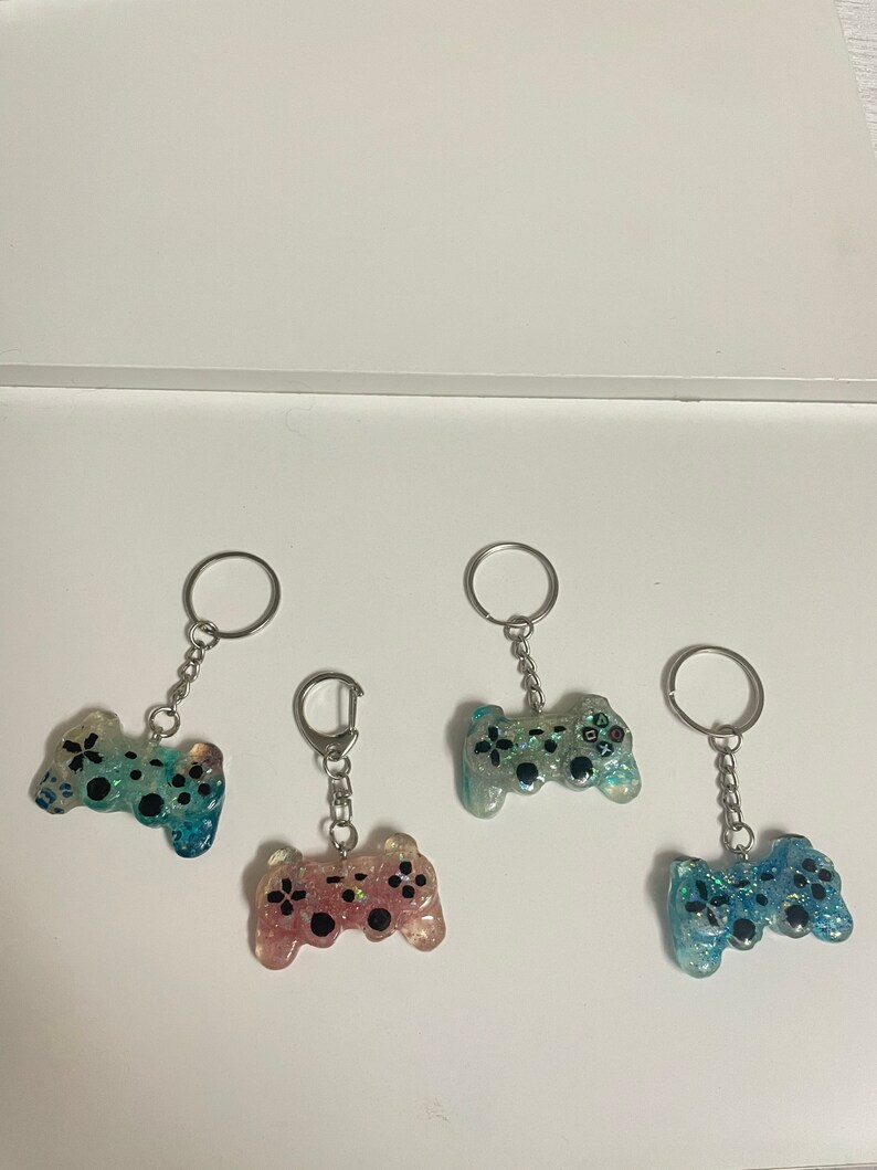 Może przedstawiać: Cztery breloczki do kluczy z żywicy w kształcie kontroler&oacute;w do gier wideo. Każdy breloczek ma srebrny pierścień i łańcuszek. Kontrolery są w odcieniach niebieskiego, r&oacute;żowego i zielonego, z czarnymi detalami guzik&oacute;w i brokatem.