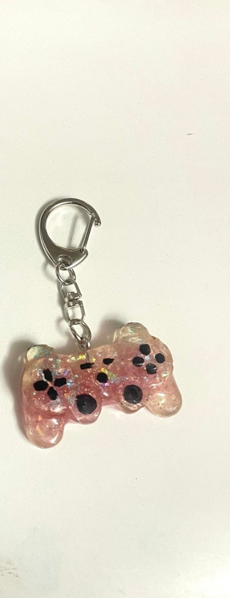 Może przedstawiać: R&oacute;żowy, błyszczący brelok z żywicy w kształcie kontrolera do gier wideo. Kontroler ma czarne przyciski oraz srebrny łańcuszek i zapięcie do mocowania do kluczy lub torby.