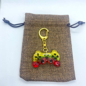 May include: A vibrant resin keychain in the shape of a video game controller. The controller has yellow, green, and red detailing and is attached to a gold-tone metal clasp. It sits on a brown drawstring bag.