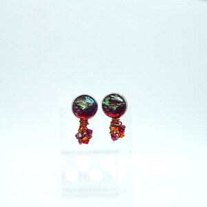 Könnte beinhalten: Ein Paar handgefertigte Ohrstecker. Jeder Ohrring hat einen runden, irisierenden Glascabochon in Lila, Grün und Rot, der in einer silbernen Lünette gefasst ist. Unterhalb des Cabochons befindet sich eine Ansammlung kleiner, facettierter Kristalle, die mit Kupferdraht umwickelt sind.