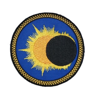 Könnte beinhalten: Ein runder gestickter Aufnäher mit goldener Umrandung. Die Mitte des Aufnähers ist blau mit einer gelben Sonne und einem schwarzen Mond, die sich überlappen.