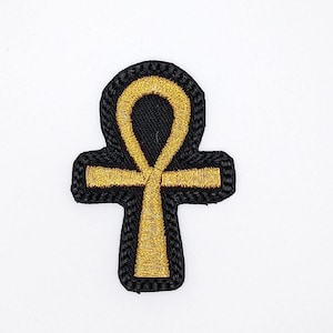 Könnte beinhalten: Gestickter Ankh-Aufnäher. Der Aufnäher hat einen schwarzen Hintergrund mit goldfarbenen Nähten. Das Ankh-Symbol, ein Kreuz mit einer Schlaufe oben, ist das Hauptdesign. Der Aufnäher hat einen schwarzen Rand und ist wahrscheinlich für Kleidung oder Accessoires.