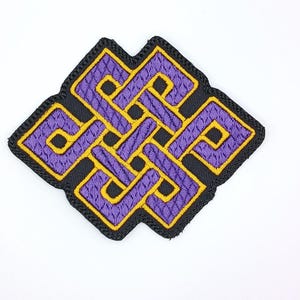 May include: Embroidered patch featuring a complex, interwoven design in purple and gold, outlined with black stitching. The patch is a square shape with rounded corners, set against a white background.
