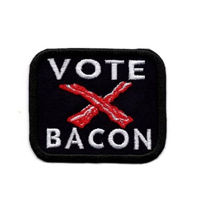 Könnte beinhalten: Ein schwarzer quadratischer Patch mit weißem Text, der "VOTE" und "BACON" sagt, mit einem roten X aus Speckscheiben in der Mitte.