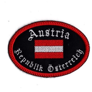 May include: An oval-shaped embroidered patch with a red border. The center of the patch is black with white text that reads "Austria Republik Österreich". A white and red Austrian flag is centered on the patch.