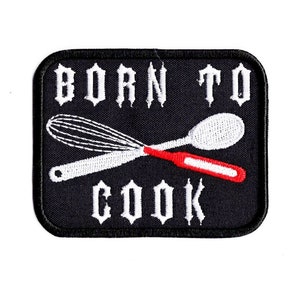 Op de afbeelding: Zwarte geborduurde patch met witte tekst die "Born to Cook" zegt en een witte garde en lepel die elkaar kruisen met een rode steel.