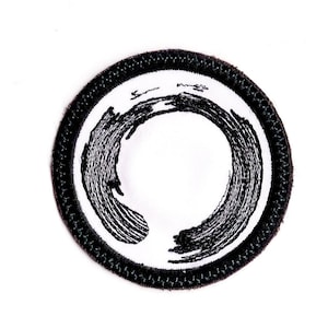May include: Black and white embroidered patch with a circle design. The circle is made up of a series of black lines that create a swirling effect.