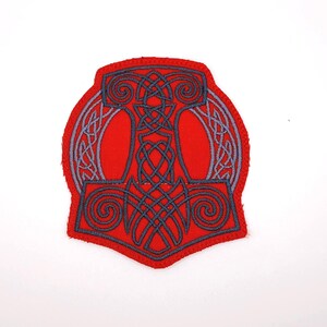 May include: A red patch featuring a gray Thor's hammer design. The hammer is intricately detailed with Celtic knot patterns and swirls. The patch has a red border and is set against a white background.