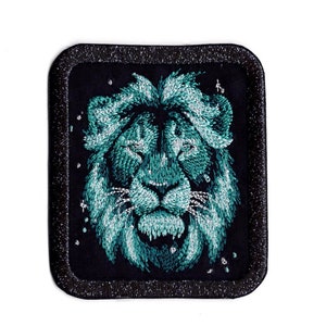 Op de afbeelding: Een gestikte turquoise leeuwenkop patch met een zwarte achtergrond en een zilveren glitterrand.