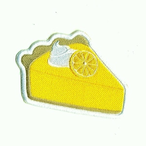 May include: A yellow lemon meringue pie with a white meringue topping and a lemon slice on top. The pie is embroidered on a white fabric.