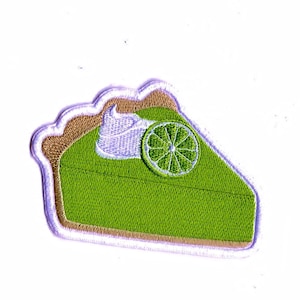 Peut inclure: Une tarte à la lime verte avec une garniture de meringue blanche et une tranche de lime sur le dessus. La tarte est brodée sur un tissu blanc.