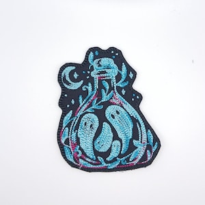Op de afbeelding: Gestikt patch met een zwarte fles met een blauwe maan en blauwe geesten erin. De fles is omringd door roze en blauwe bloemmotieven.