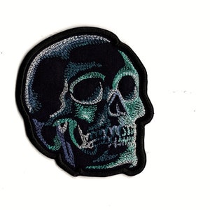 Peut inclure: Un écusson brodé noir en forme de crâne avec des détails verts et bleus. Le crâne est tourné vers la droite et présente un design détaillé.