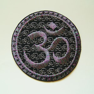 May include: A black circular patch with a silver Om symbol embroidered in the center. The background of the patch is a repeating pattern of small squares.