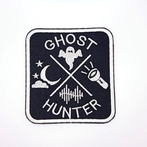 May include: Embroidered patch with a black background and white border. The patch features the words "GHOST HUNTER" in white, along with a ghost, moon, stars, flashlight, and sound wave design. The patch is square with rounded corners.