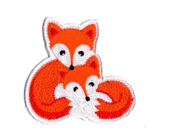 Op de afbeelding: Een geborduurde patch met twee vossen, een grotere en een kleinere, beide met feloranje vacht, witte borst- en gezichtsmarkeringen en zwarte ogen en neuzen. De vossen zijn samen afgebeeld waarbij de kleinere tegen de grotere aanligt, alles afgewerkt met een witte rand.