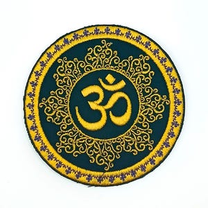 May include: Round embroidered patch featuring the Om symbol in gold thread on a dark teal background. The symbol is surrounded by ornate gold filigree and a border of small purple flowers. The patch is circular.