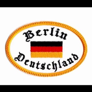 May include: An oval-shaped embroidered patch with a yellow border. The patch features the text "Berlin Deutschland" in black cursive lettering and a German flag in the center.