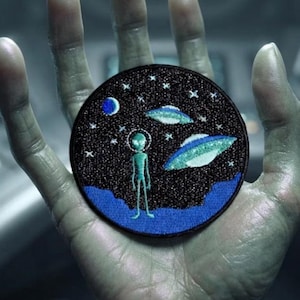 Peut inclure: Un écusson brodé circulaire représentant une scène spatiale avec un extraterrestre vert clair debout sur une planète bleue. Le fond est noir avec des étoiles blanches, une lune bleue et deux soucoupes volantes dans des tons de vert et de bleu.