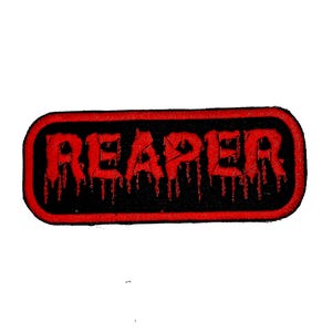 May include: A rectangular black patch with a red border. The word "REAPER" is embroidered in red, with a dripping blood effect. The patch is likely for clothing or accessories.