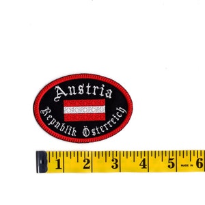 May include: An oval-shaped embroidered patch with a black background and white text that reads "Austria Republik &Ouml;sterreich". The patch features the Austrian flag in the center, with red and white stripes and a white emblem.