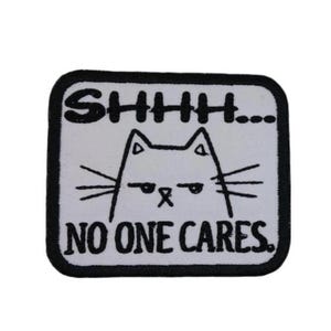 May include: A white square patch with black embroidered text and a cartoon cat. The text reads "SHHHH... NO ONE CARES."