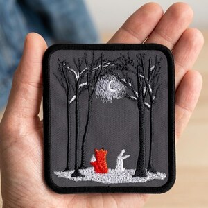 May include: Embroidered patch featuring a forest scene with a red fox and a white rabbit under a moonlit sky. The patch has a dark gray background with black trees and a black border. The patch is rectangular with rounded corners.