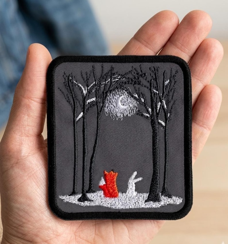 May include: Embroidered patch featuring a forest scene with a red fox and a white rabbit under a moonlit sky. The patch has a dark gray background with black trees and a black border. The patch is rectangular with rounded corners.