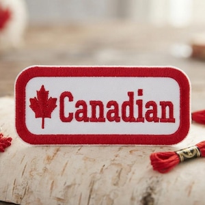 Könnte beinhalten: Ein rechteckiger gestickter Aufnäher mit weißem Hintergrund und rotem Rand. Der Aufnäher zeigt links ein rotes Ahornblatt-Symbol und rechts das Wort "Canadian", das in roten Groß- und Kleinbuchstaben eingestickt ist.